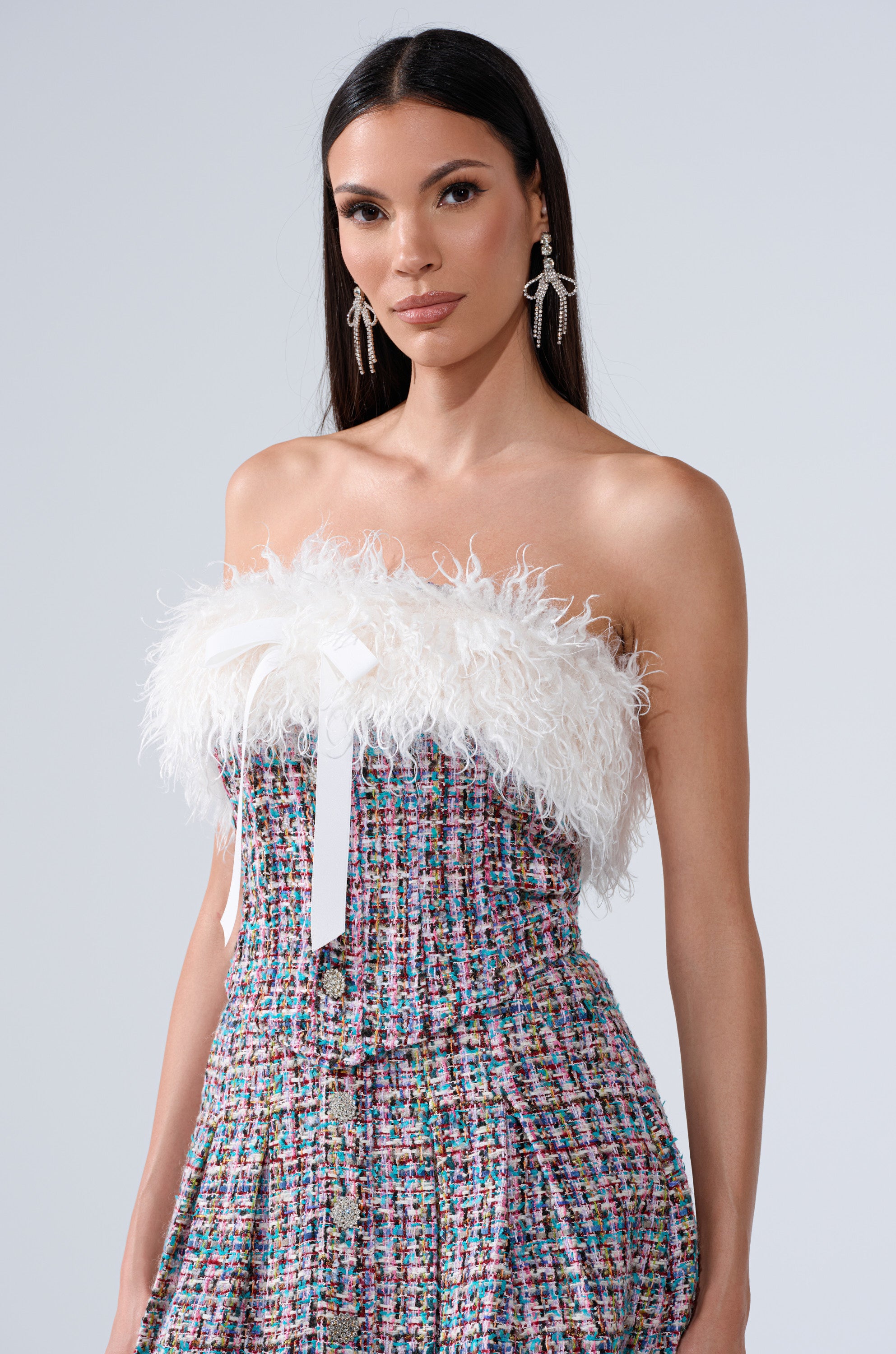 A woman with long dark hair wears the FUR TRIM TWEED CORSET, featuring a feathered faux fur top and colorful tweed fabric. She accessorizes with silver dangling earrings against a light gray background.