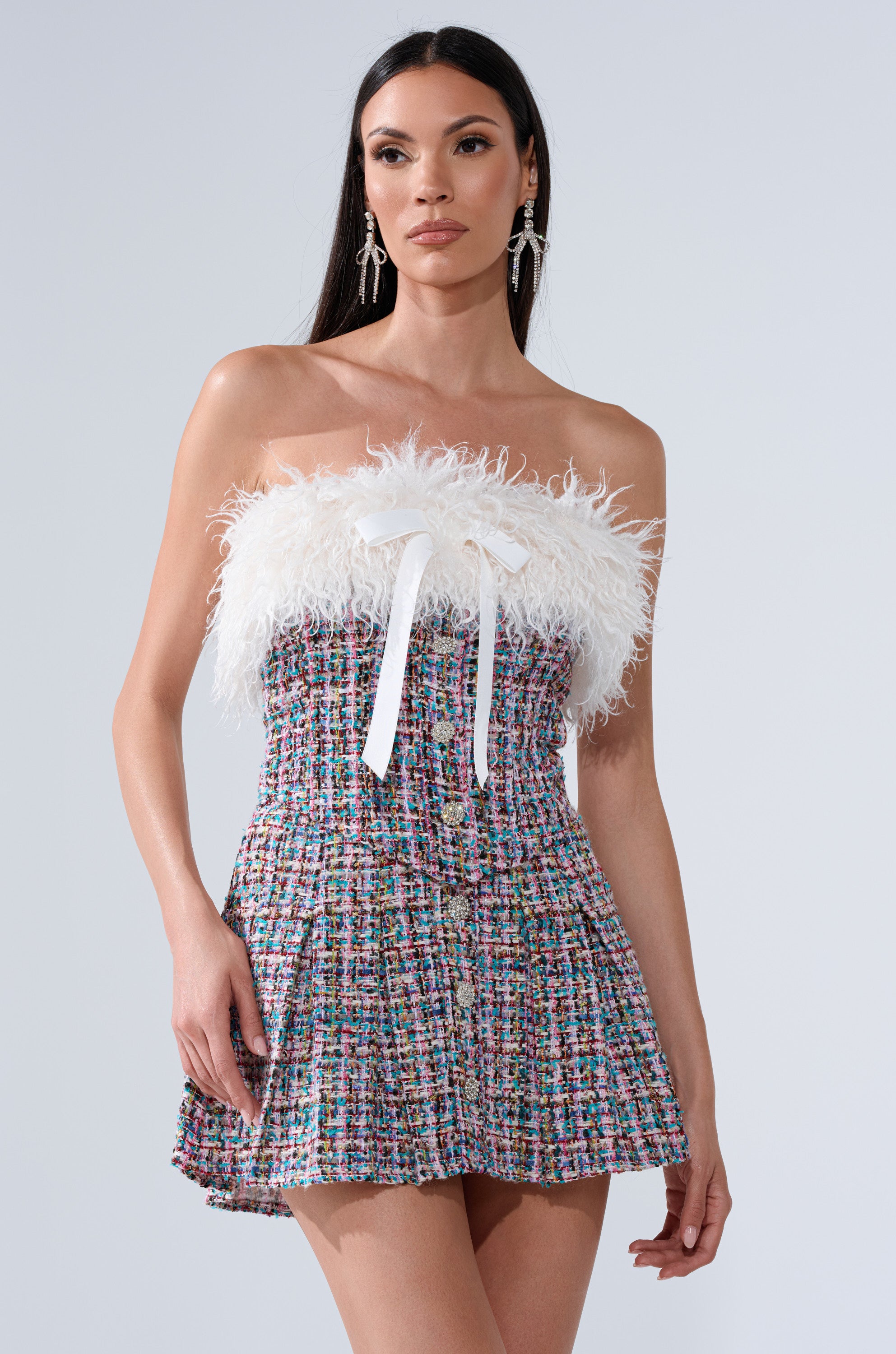 A woman models the FUR TRIM TWEED CORSET, a strapless multicolored tweed mini dress with white feathered faux fur trim and a ribbon bow on the bodice, against a plain background. She has long straight hair and rhinestone earrings.