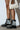 A person stands against a white wall wearing AZALEA WANG TWINKLEY black pearl embellished combat booties. They hold a small white handbag with pearl accents, perfectly enhancing the minimalist aesthetic of the setting.