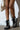 A person stands against a white wall, holding a pearl-embellished handbag. They stylishly pair their short outfit with AZALEA WANG TWINKLEY BLACK PEARL EMBELLISHED COMBAT BOOTIE in faux leather, featuring studded accents.