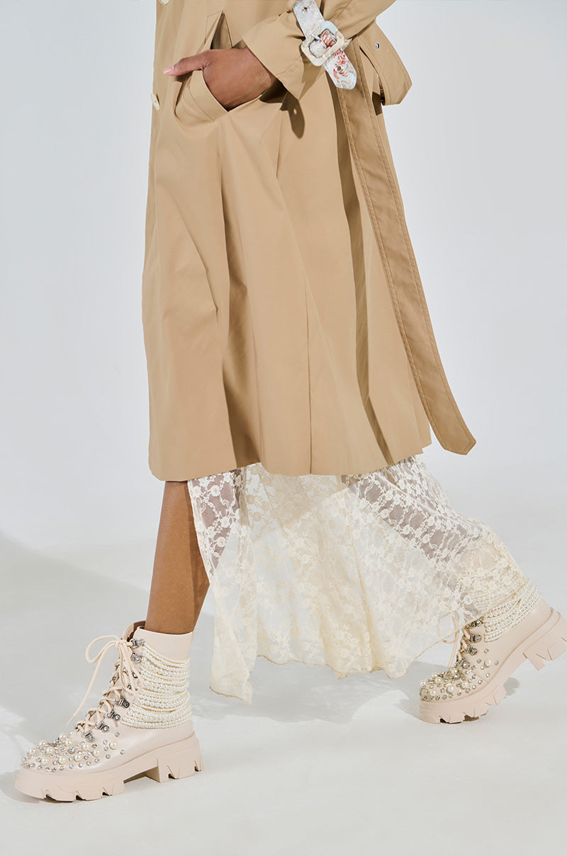 A person strides confidently in AZALEA WANG TWINKLEY Bone Pearl Embellished Combat Booties, paired with a beige trench coat and white lace skirt against an off-white backdrop. 2