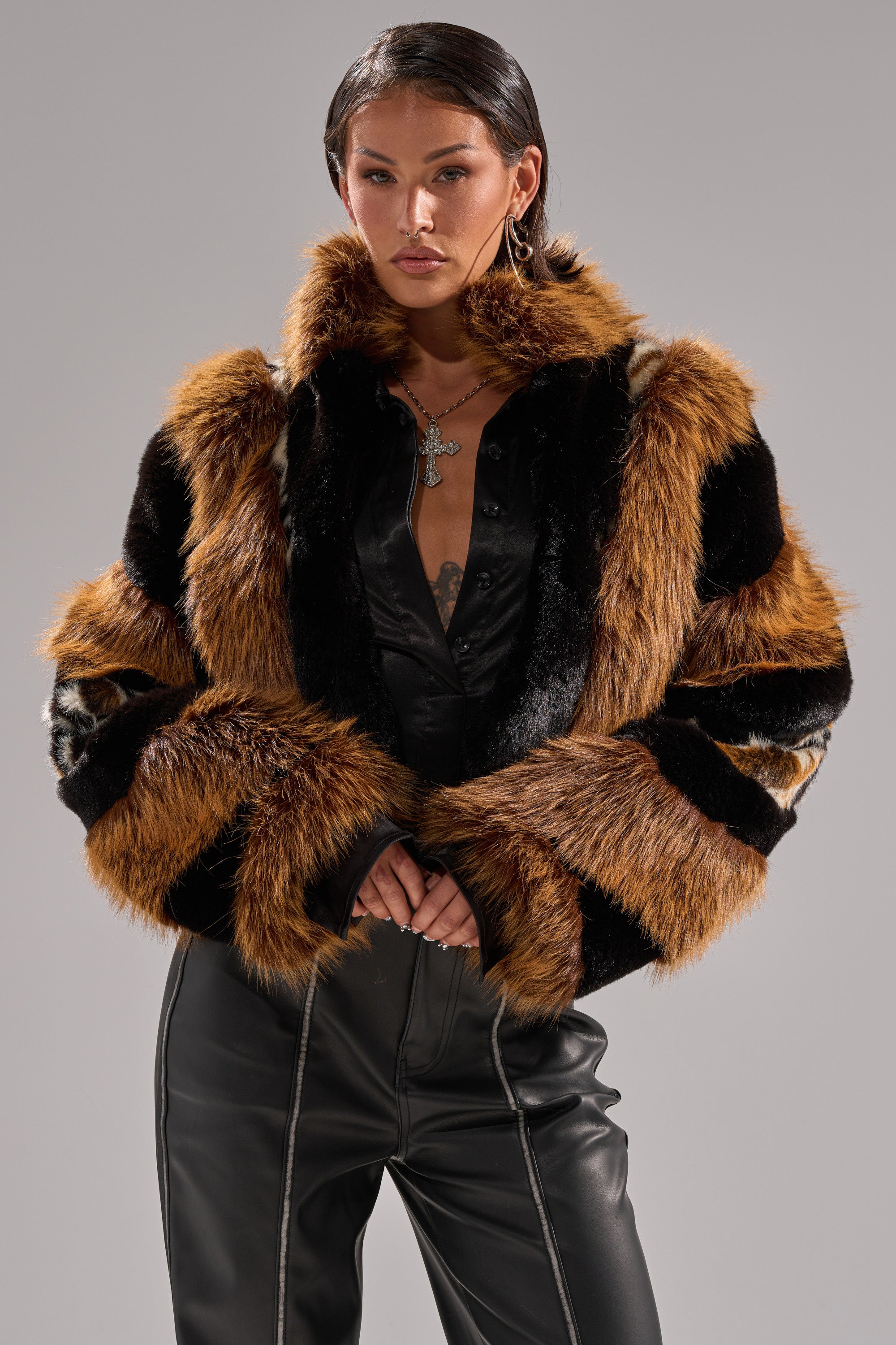 A woman stands confidently against a plain gray background, wearing the TWISTER PATCHWORK FAUX FUR jacket in brown, black, and white over a black satin top and black leather pants.