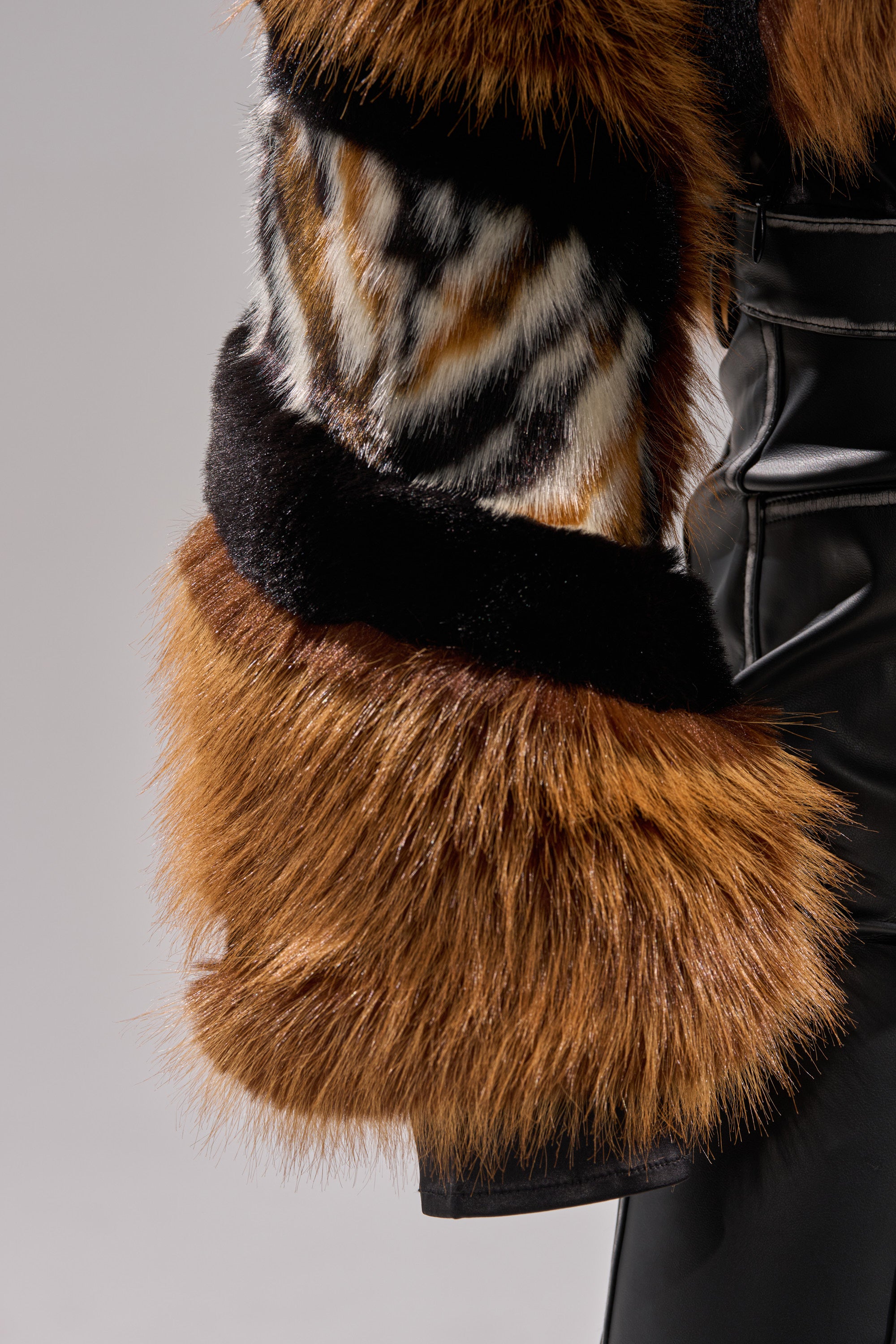 Close-up of a person's arm wearing the TWISTER PATCHWORK FAUX FUR jacket with a tiger-like pattern and brown trim, paired with black leather pants, set against a plain gray background.