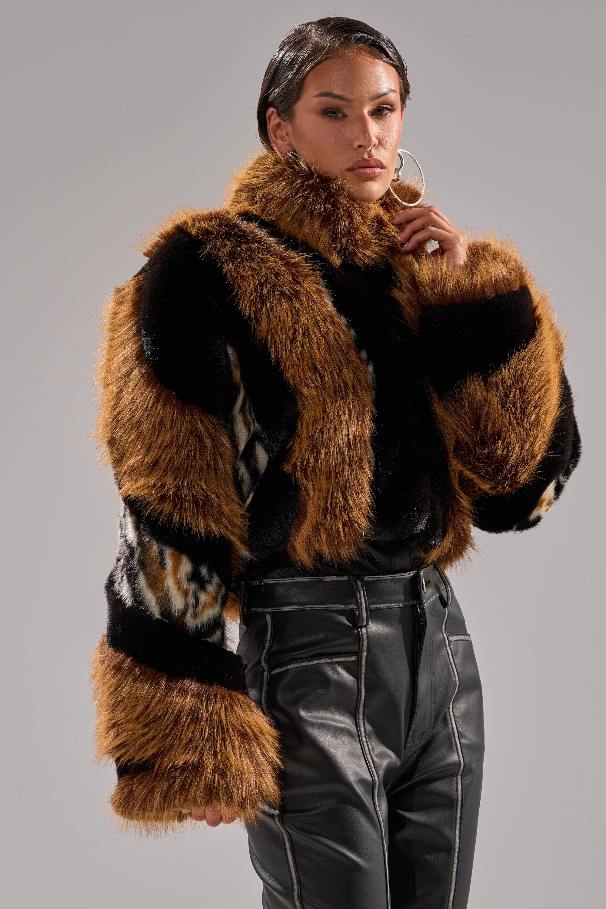 A woman with slicked-back hair wears large hoop earrings, high-waisted black leather pants, and the TWISTER PATCHWORK FAUX FUR jacket featuring brown, black, and animal print patches, posing against a plain gray background.