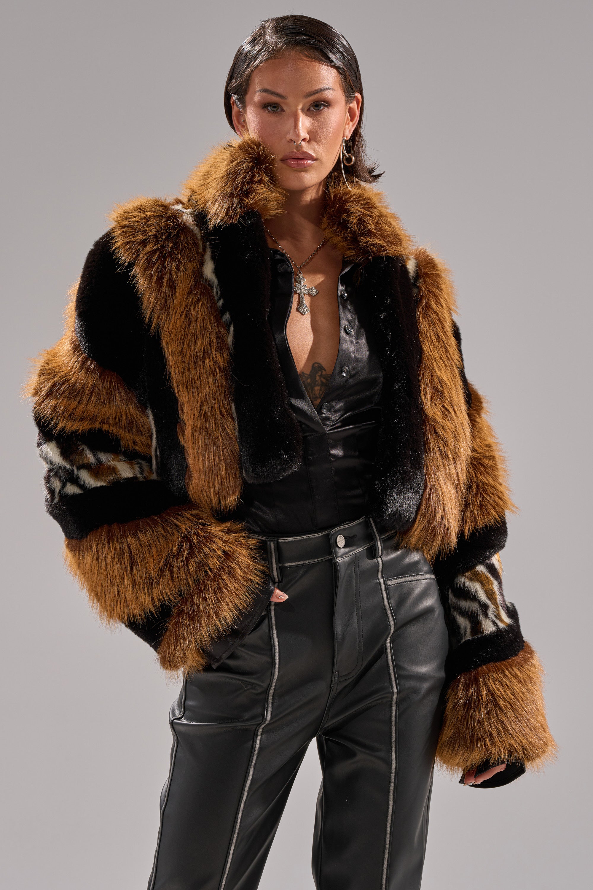 A woman with short dark hair confidently poses against a gray background in the bold TWISTER PATCHWORK FAUX FUR jacket with brown, black, and white patterns, a black satin shirt, and high-waisted black leather pants with white stitching.
