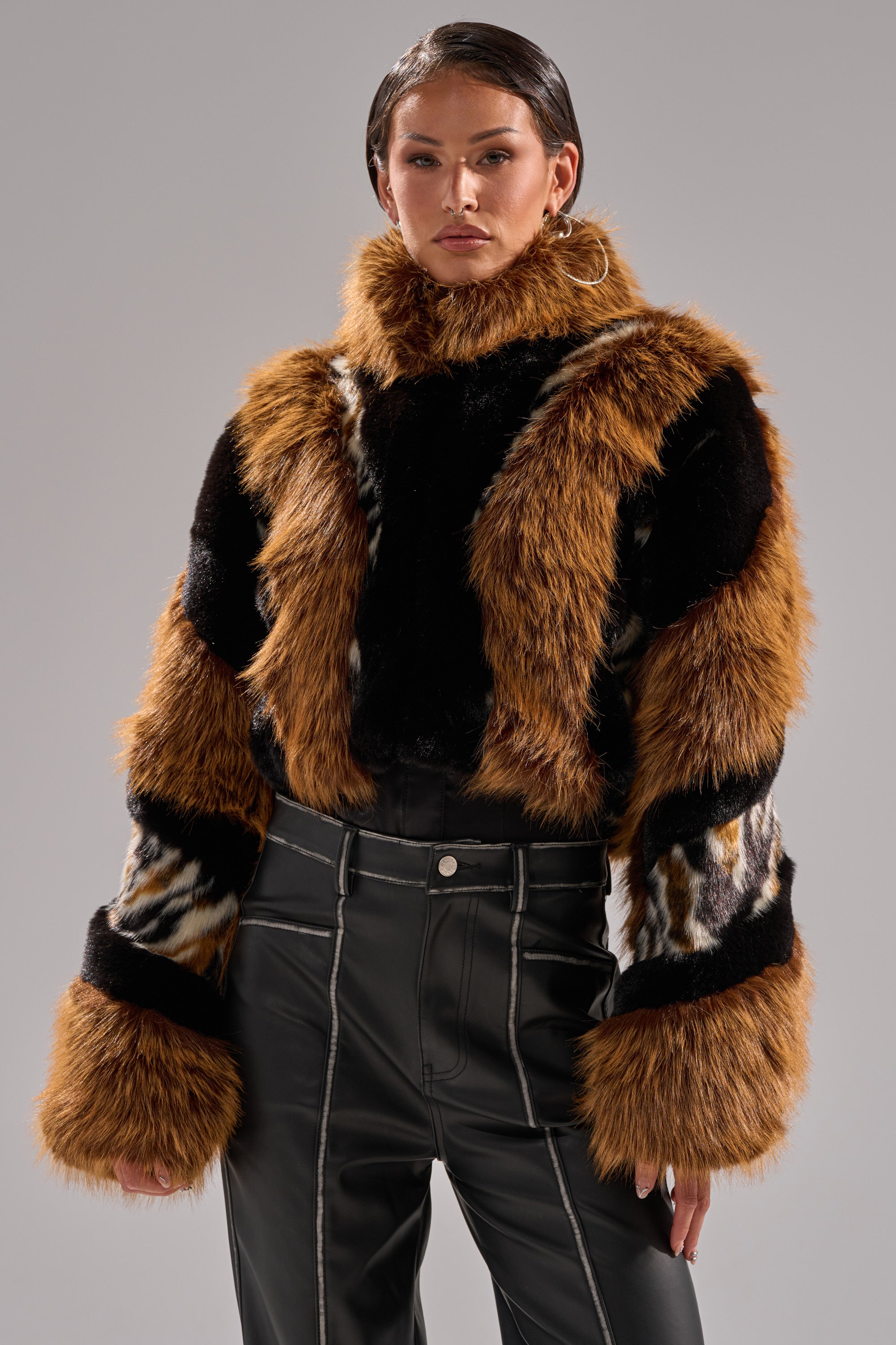 A woman stands confidently in the TWISTER PATCHWORK FAUX FUR jacket featuring brown, black, and white patterns, paired with high-waisted black pants with white stitching, against a plain gray background.