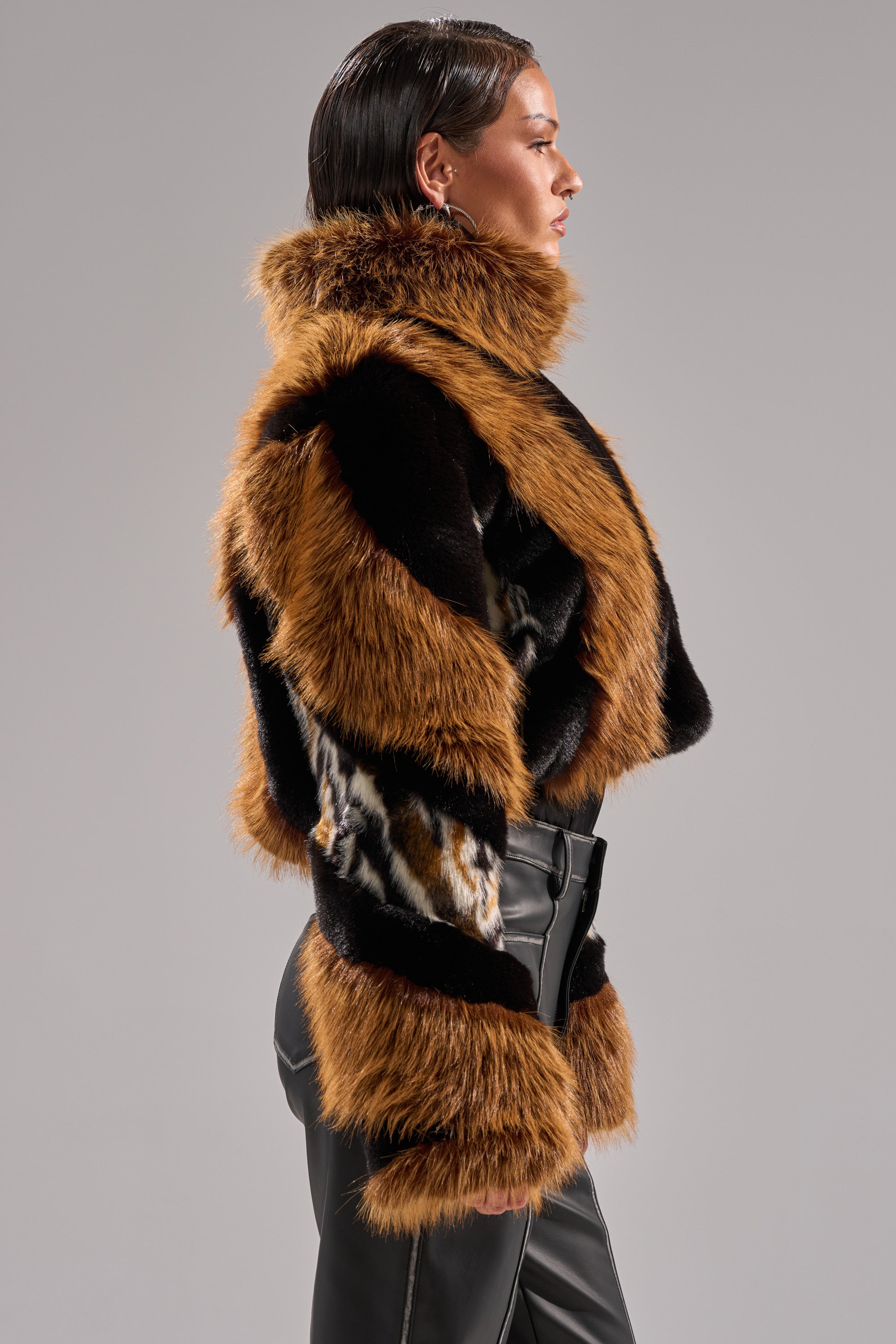A person with slicked-back hair wears the TWISTER PATCHWORK FAUX FUR jacket with brown, black, and animal print designs, paired with high-waisted black leather pants, standing in profile against a plain gray background.
