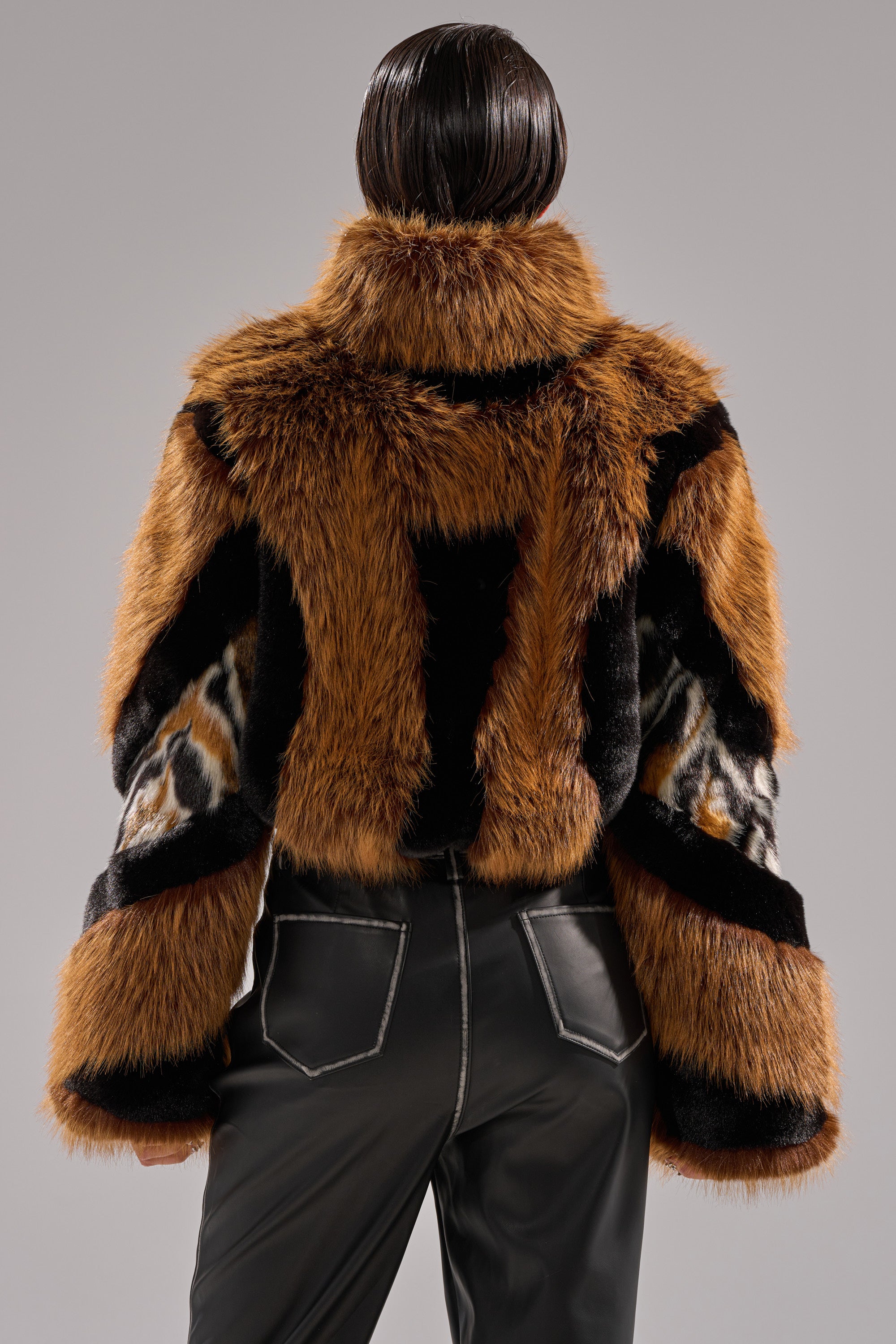 Wearing the TWISTER PATCHWORK FAUX FUR jacket in bold brown, black, and white patterns, a person with slicked-back dark hair is shown from the back, paired with black pants featuring white stitching against a plain background.