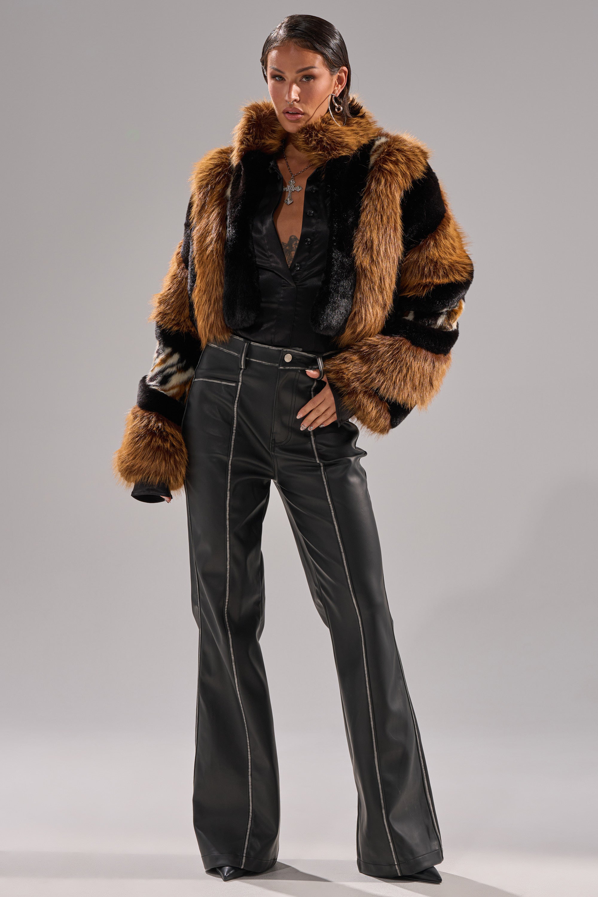 A woman exudes confidence in the TWISTER PATCHWORK FAUX FUR jacket with brown, black, and tan stripes, paired with a black buttoned top and high-waisted black leather pants. Slicked-back hair and bold jewelry finish her striking look.