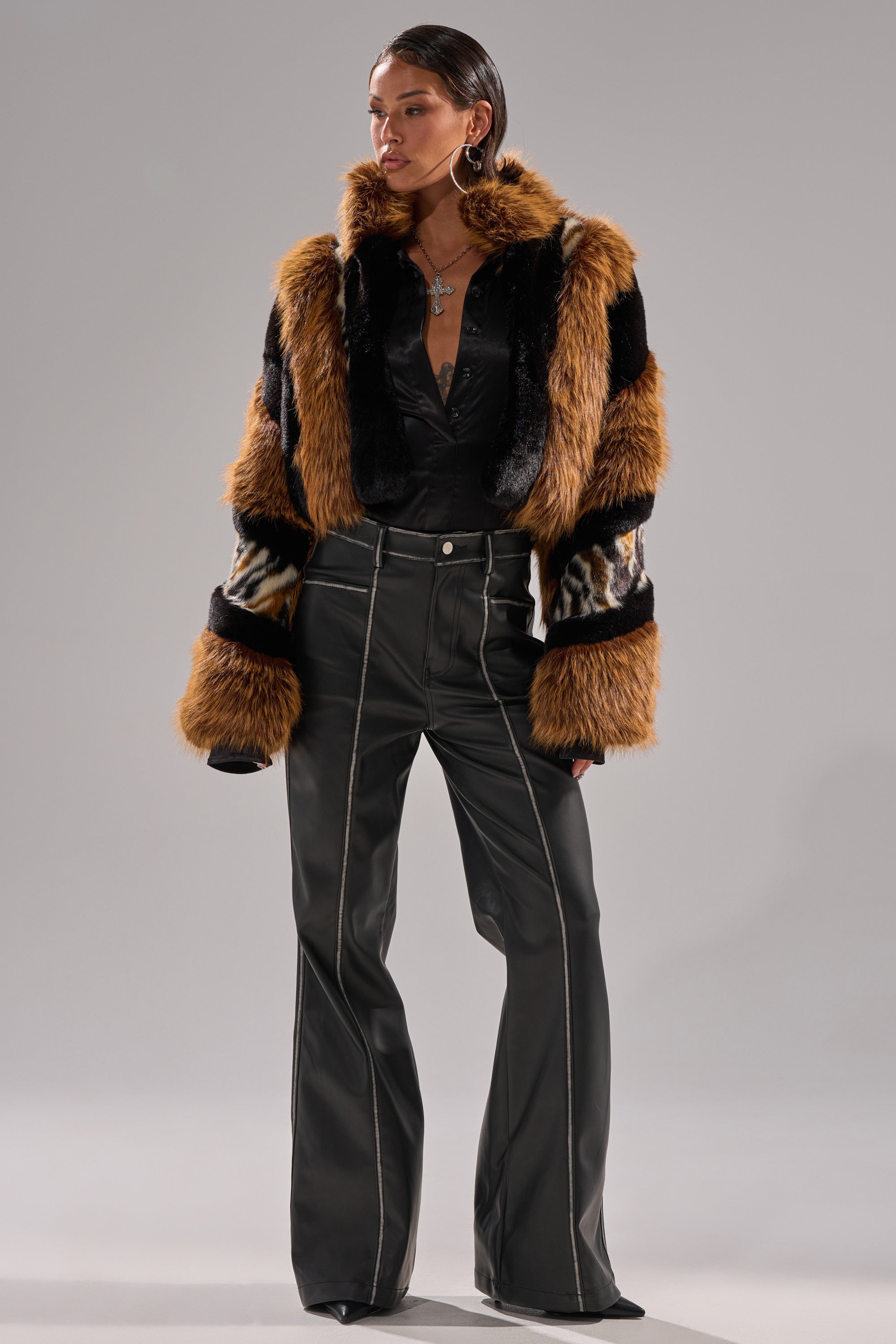 A person stands confidently in black flared pants and a top, layering on the TWISTER PATCHWORK FAUX FUR jacket in brown, black, and cream. Large earrings and layered necklaces complete their striking, modern look.