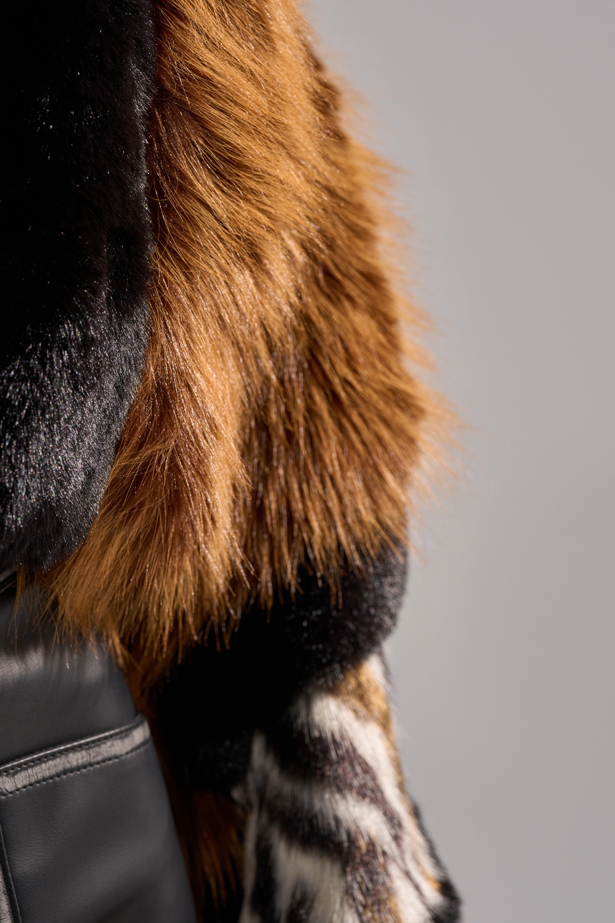 Close-up of a person wearing a black leather garment with the TWISTER PATCHWORK FAUX FUR stole, featuring dark brown, reddish, and black faux fur textures draped over their shoulder against a neutral background.
