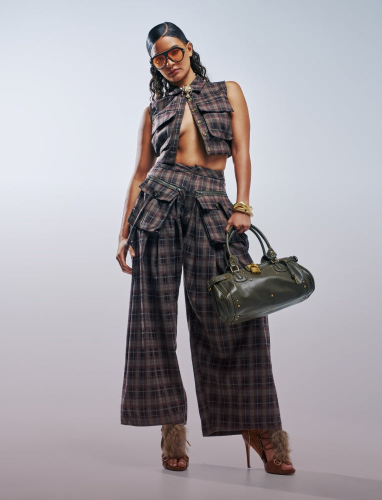A model wearing a matching plaid crop top and wide-leg pants set, holding an olive green handbag, photographed against a light gray studio background.