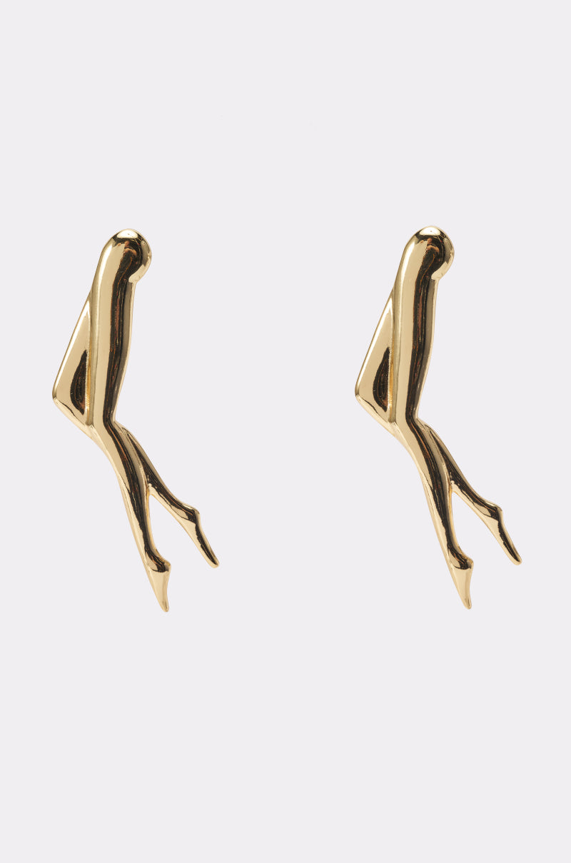 SHOW YA LEGS EARRINGS are gold metallic statement earrings shaped like abstract human legs, each with a bent knee and pointed toes, making a bold fashion statement on any plain background.
