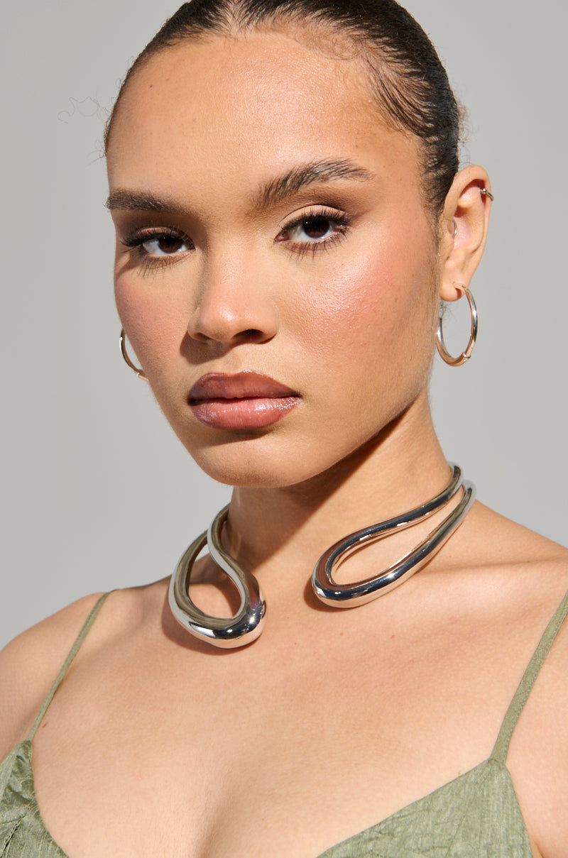 A person with hoop earrings and an ASH ABSTRACT NECKLACE poses against a neutral background. They wear a green sleeveless top, and their hair is neatly pulled back. 2