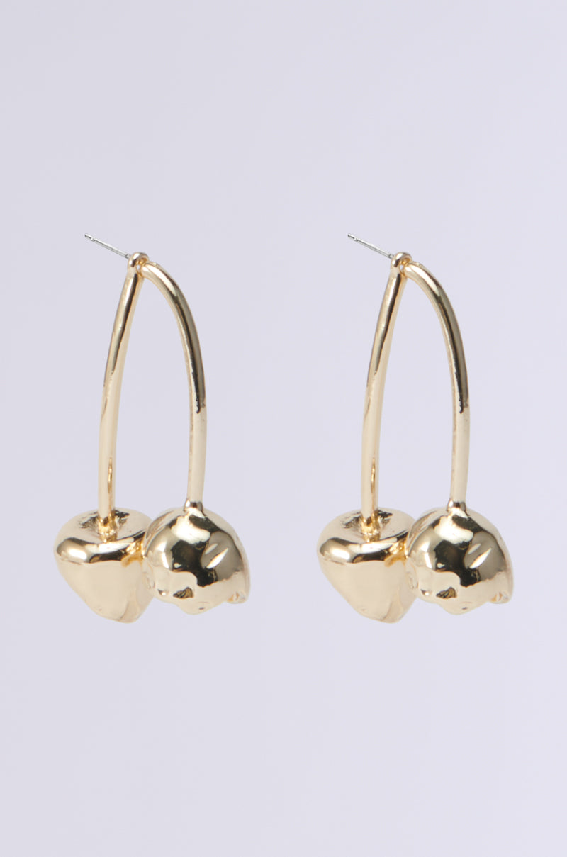 Gold earrings featuring a distinctive design that mimics hollow cherries are set against a pale backdrop. Known as the CHERRY BOMB EARRING, each piece showcases two glossy, spherical elements connected by a slender, curved faux gold stem.