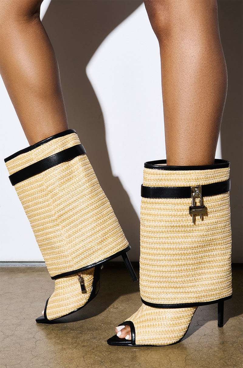 The AZALEA WANG TYLOR BEIGE WOVEN BOOTIE displays a unique woven raffia texture with black accents and gold locks on high-heeled, knee-high boots. These elegant stiletto heels stand gracefully on a glossy beige floor.