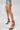 A person in frayed denim shorts and AZALEA WANG TZILLA MULTI COLOR ROPE STILETTO SANDALS holds a small white woven handbag while standing against a plain, light background.