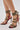 A close-up of a person wearing the AZALEA WANG TZILLA MULTI COLOR ROPE STILETTO SANDAL, featuring colorful braided straps wrapping around the ankles and feet, and stiletto heels with a bold red and black marbled pattern.