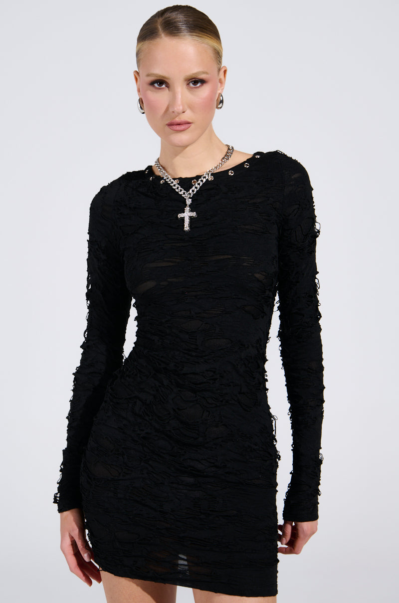 In a studio, someone models the LOTS OF LOVE LACE MINI DRESS featuring a curve-hugging silhouette and long sleeves. Their short, slicked-back hair complements their silver cross necklace and earrings. The background is plain and light-colored.