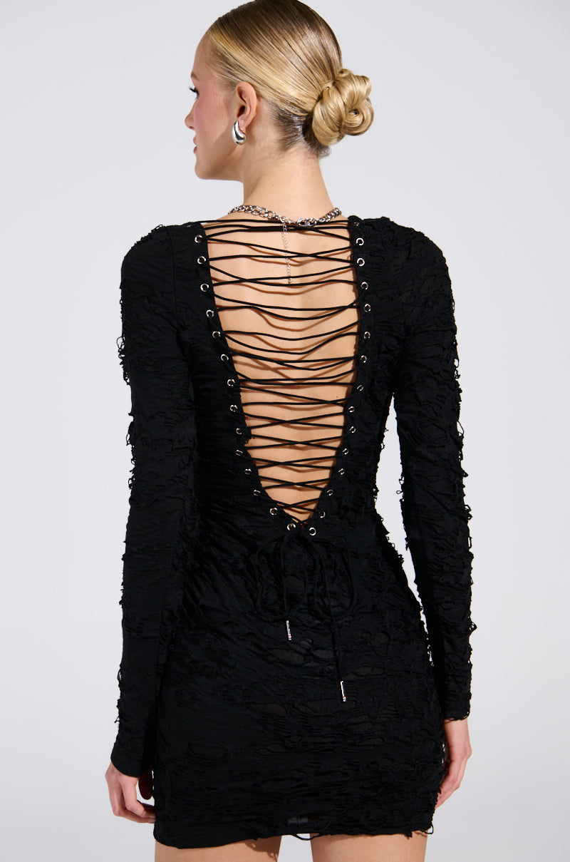 A person is wearing the LOTS OF LOVE LACE MINI DRESS, a curve-hugging LBD with long sleeves and a deep lace-up V-back design featuring eyelets and a criss-cross pattern. They have blonde hair in a bun, accessorized with earrings and a necklace.