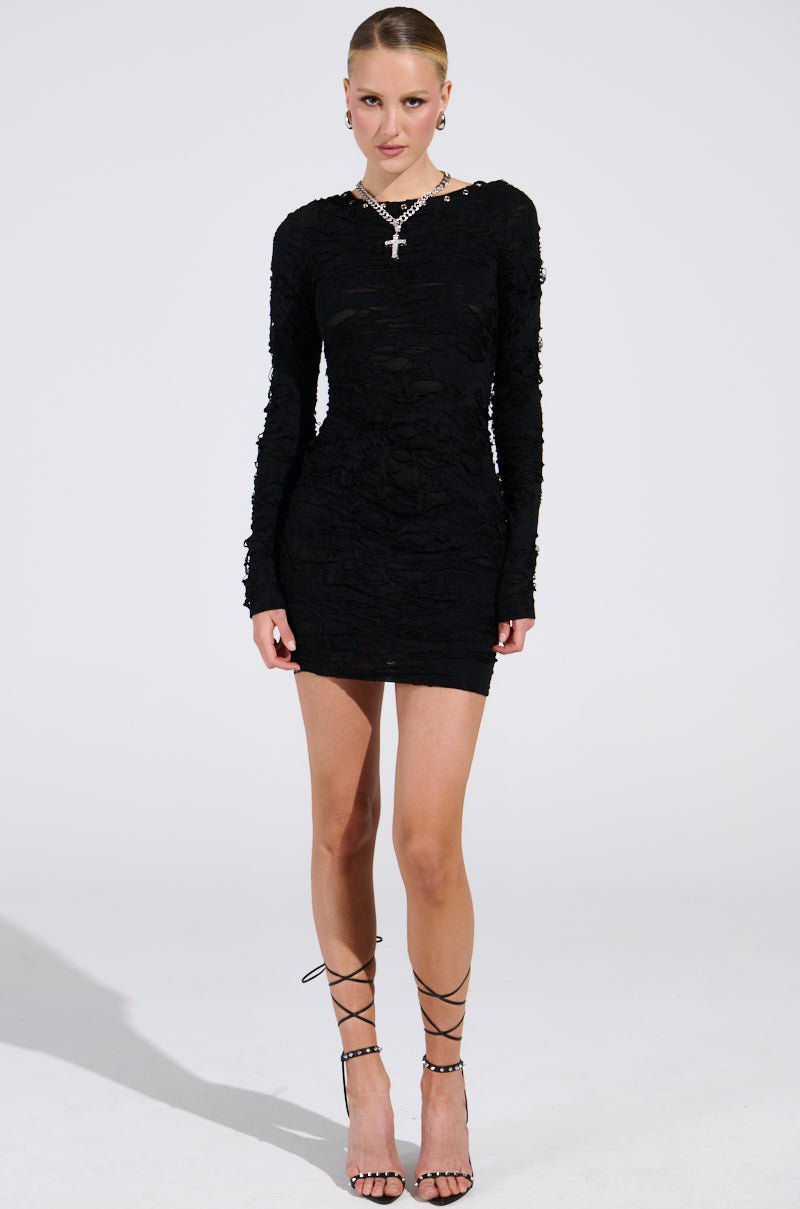 A person in a LOTS OF LOVE LACE MINI DRESS with a curve-hugging silhouette stands against a plain background. They're wearing strappy high-heeled sandals, a cross pendant necklace, and have sleekly styled hair.