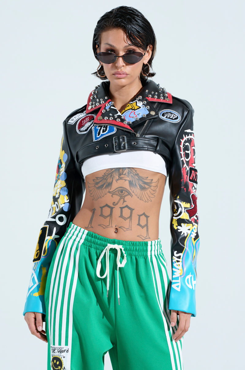 Wearing a punk-inspired PHOENIX ULTRA CROP MOTO jacket with sunglasses and colorful studs, this individual pairs green track pants and flaunts a tattooed midriff featuring "1999" against a plain, light backdrop.