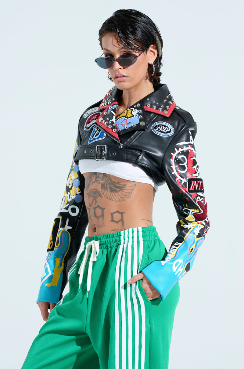 A person flaunts a punk-inspired look with the colorful PHOENIX ULTRA CROP MOTO jacket, paired with green Adidas track pants. Sporty sunglasses, wet-look hair, and visible torso tattoos complete their edgy and modern style.