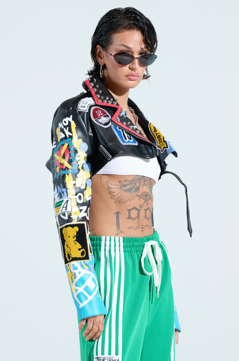 Someone in sunglasses, a PHOENIX ULTRA CROP MOTO jacket with a punk-inspired design, a white crop top, and green drawstring pants. Tattoos adorn their torso as they stand against a light background.