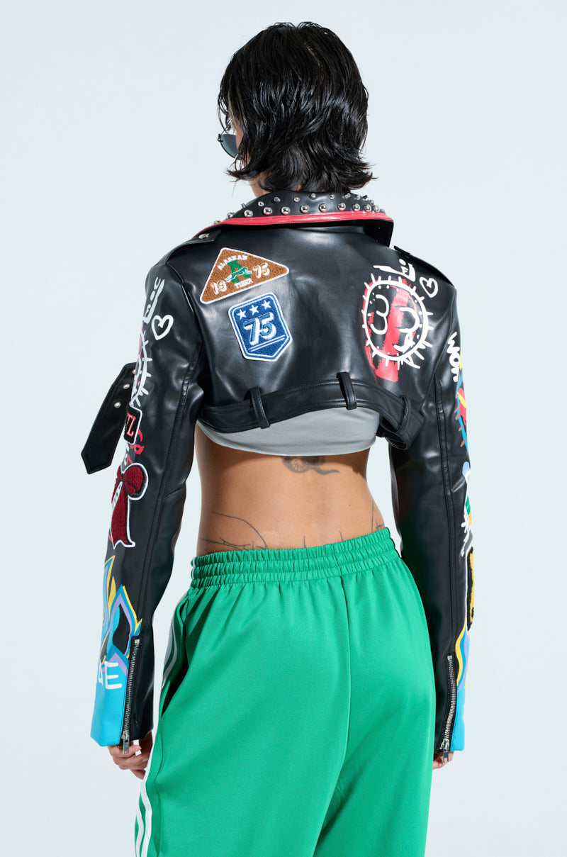 A person is facing away, donning the PHOENIX ULTRA CROP MOTO jacket in black faux leather with colorful patches, studs, and punk designs. Their gray top peeks out from the cropped silhouette, paired with green sweatpants with white stripes. They sport short dark hair to complete the look.