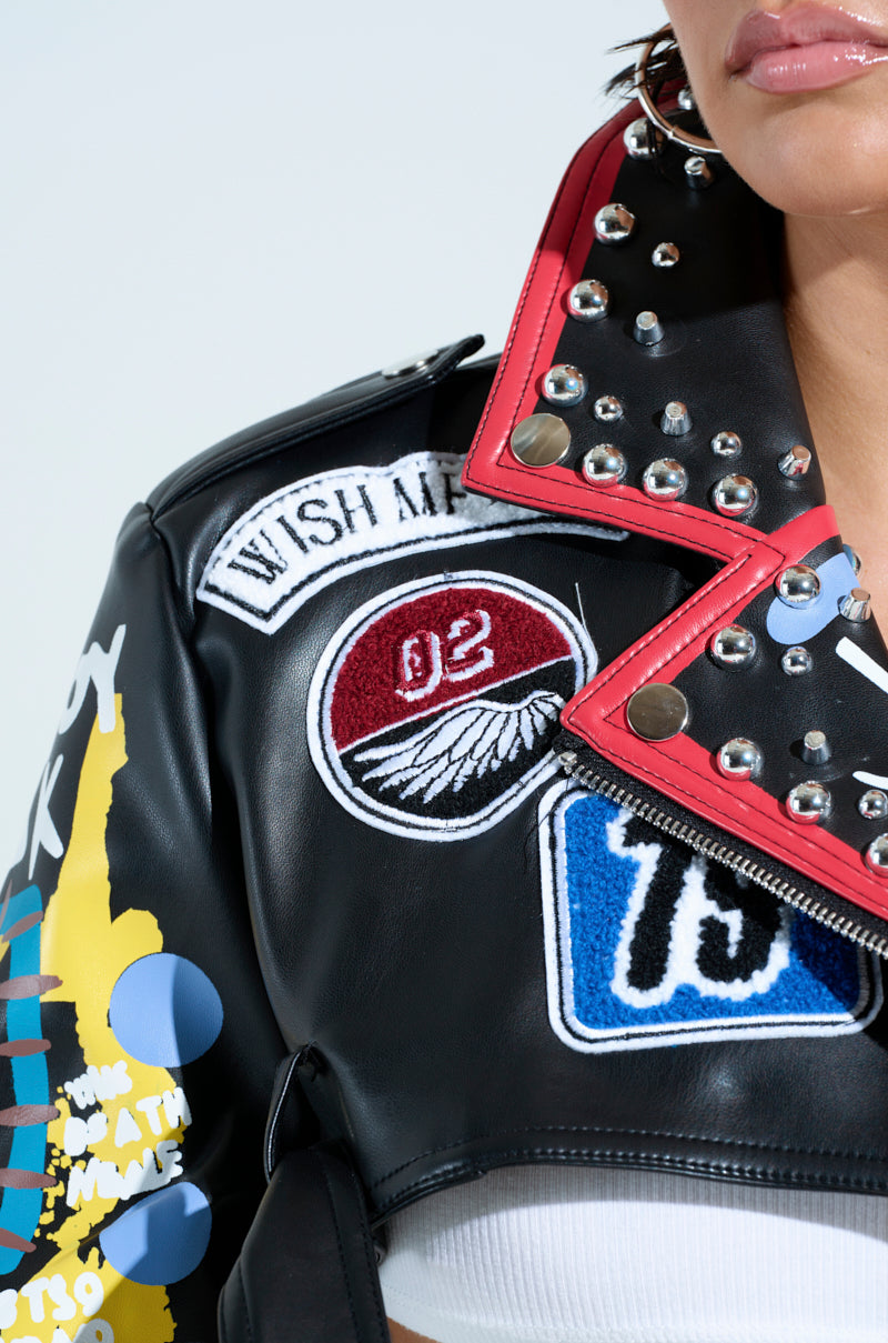 A close-up of someone wearing the PHOENIX ULTRA CROP MOTO, a punk-inspired faux leather jacket adorned with colorful patches and studded details. Featuring patches with "02" and "Wish Me," along with red trim and silver studs on the collar for added edge to its cropped silhouette.