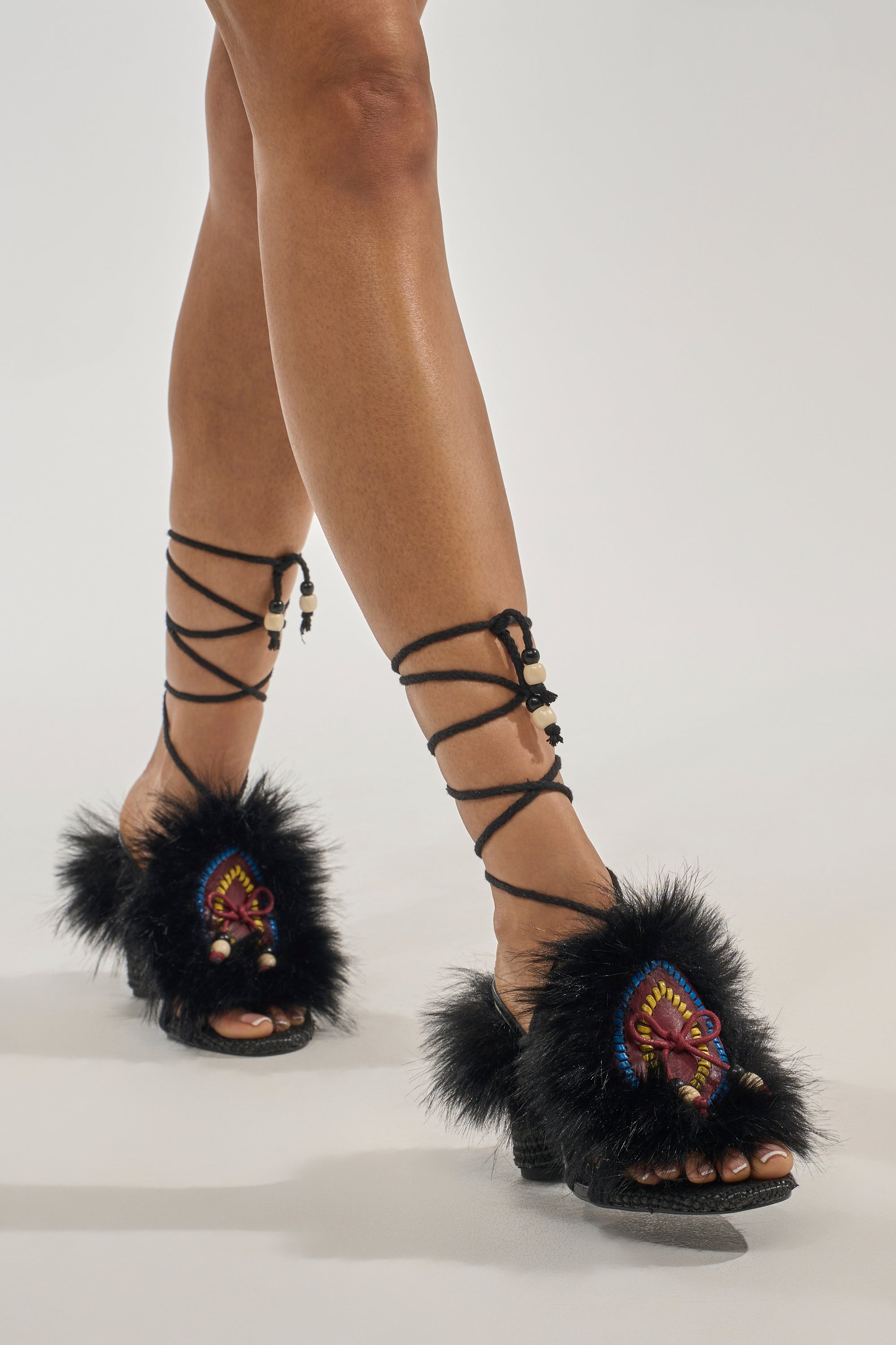 A person wears AKIRA GOLD UMIKA BLACK FUR BLOCK SANDALS with black faux fur, colorful embroidered patches, beads, and black laces wrapping around the lower legs. The background is plain and light colored. 0