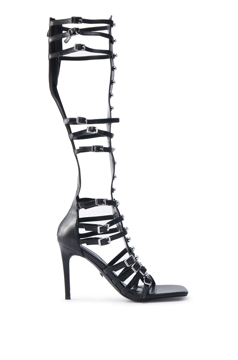 The AZALEA WANG UNDERWOOD BLACK GLADIATOR STILETTO SANDAL is a knee-high faux leather sandal with multiple horizontal straps, silver buckles, and decorative studs along the front for a bold statement look.
