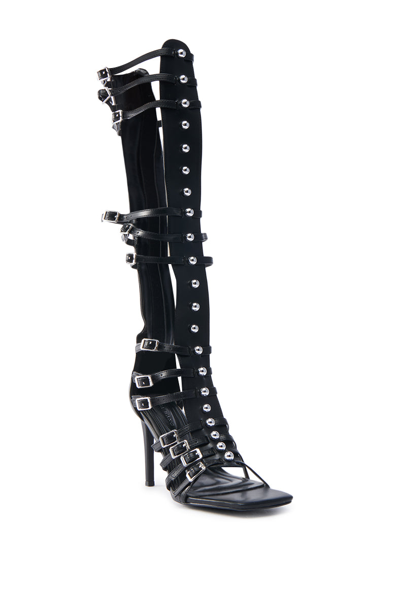 The AZALEA WANG UNDERWOOD BLACK GLADIATOR STILETTO SANDAL is a knee-high faux leather shoe with multiple buckled straps and silver grommet accents running vertically up the front.