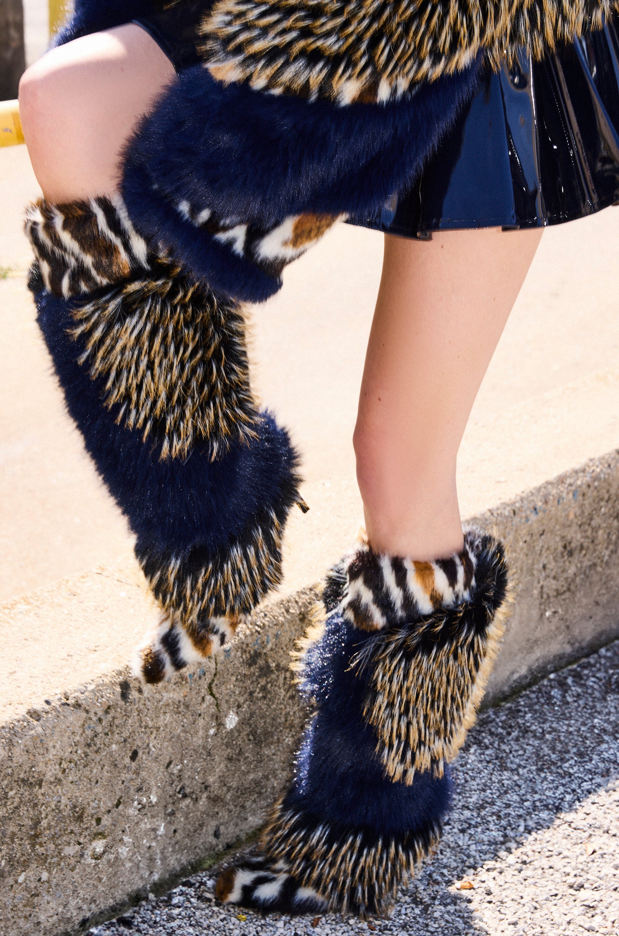 A person in a shiny black skirt and AZALEA WANG UPENDO NAVY MULTI FUR BOOTS with blue, black, white, and brown animal print stands on a concrete sidewalk.