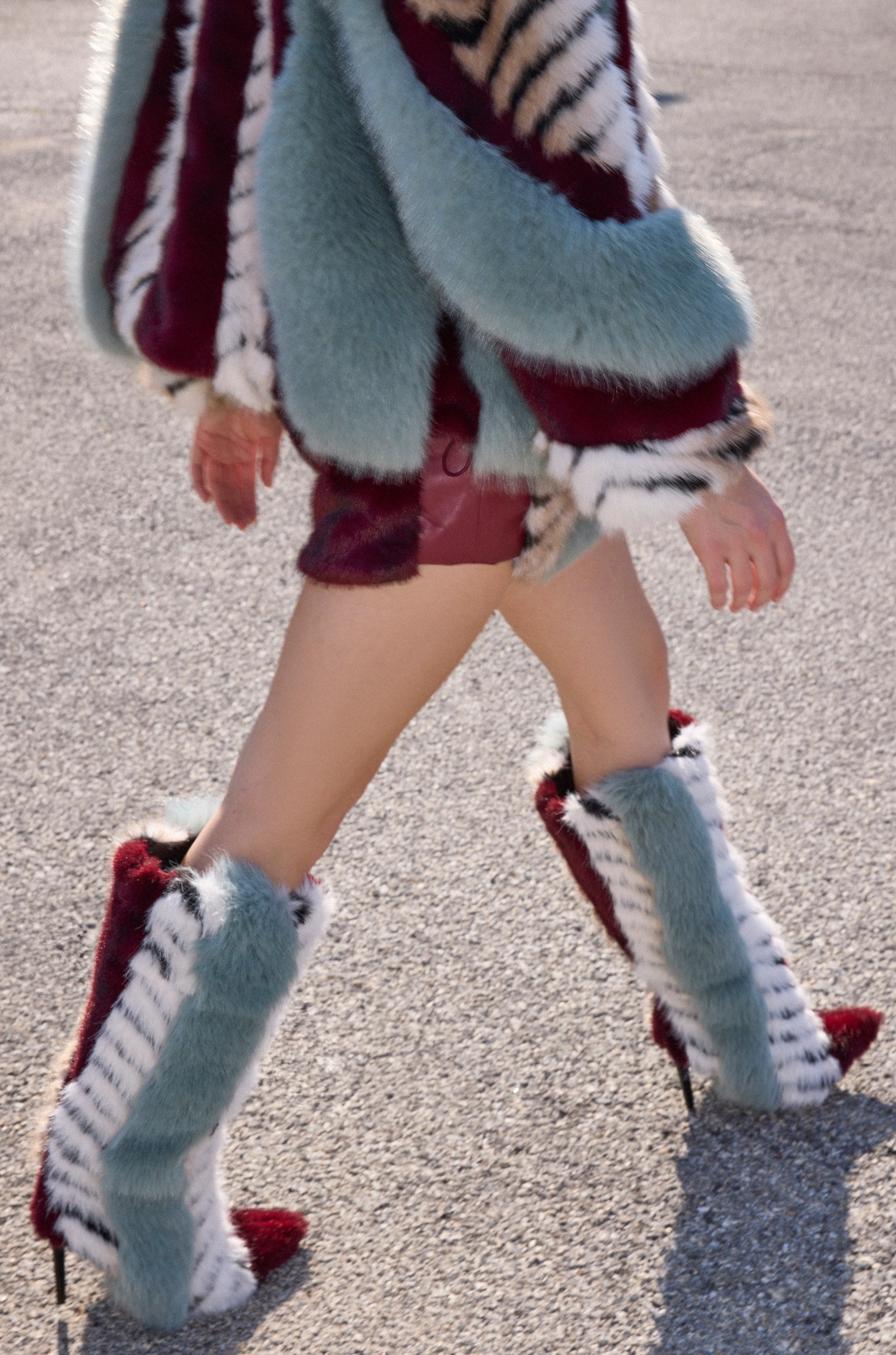 A person walks on pavement in a colorful faux fur coat, showing bare legs and wearing the AZALEA WANG UPSELLA BLUE MULTI STILETTO BOOT—knee-high boots in bold shades of blue, burgundy, and white.