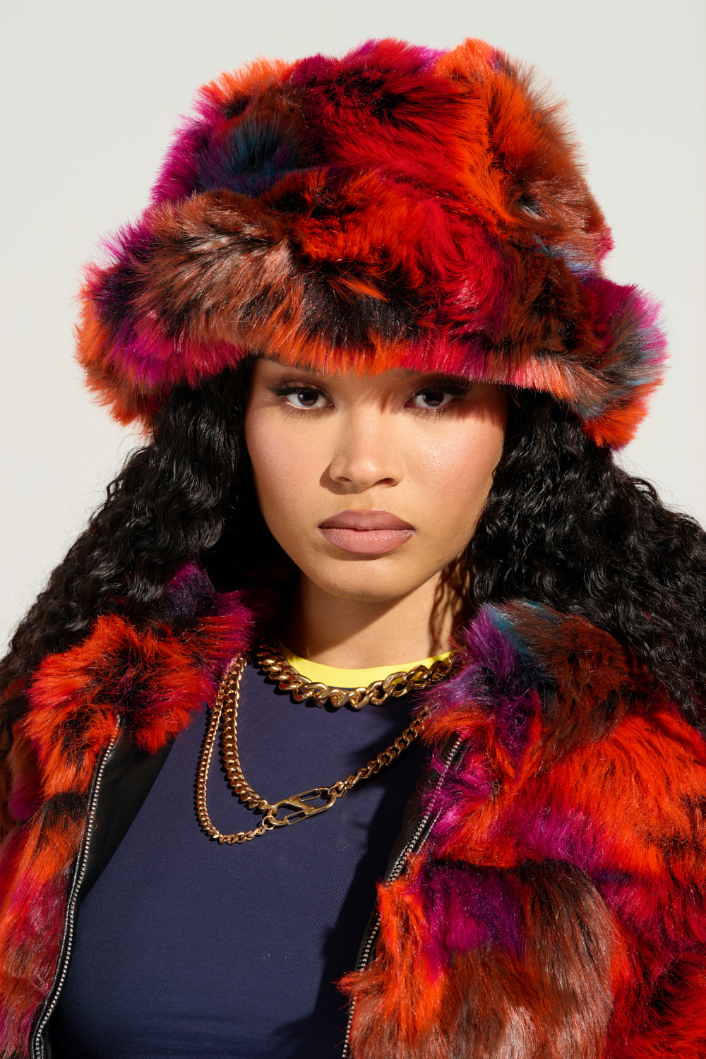 Wearing the bold UPSETTER FAUX FUR BUCKET HAT, a person sports a vibrant jacket in orange, red, and purple hues. They pair it with a navy top and layered gold chains. Their long curly hair frames their face as they gaze directly at the camera against a neutral backdrop. 0