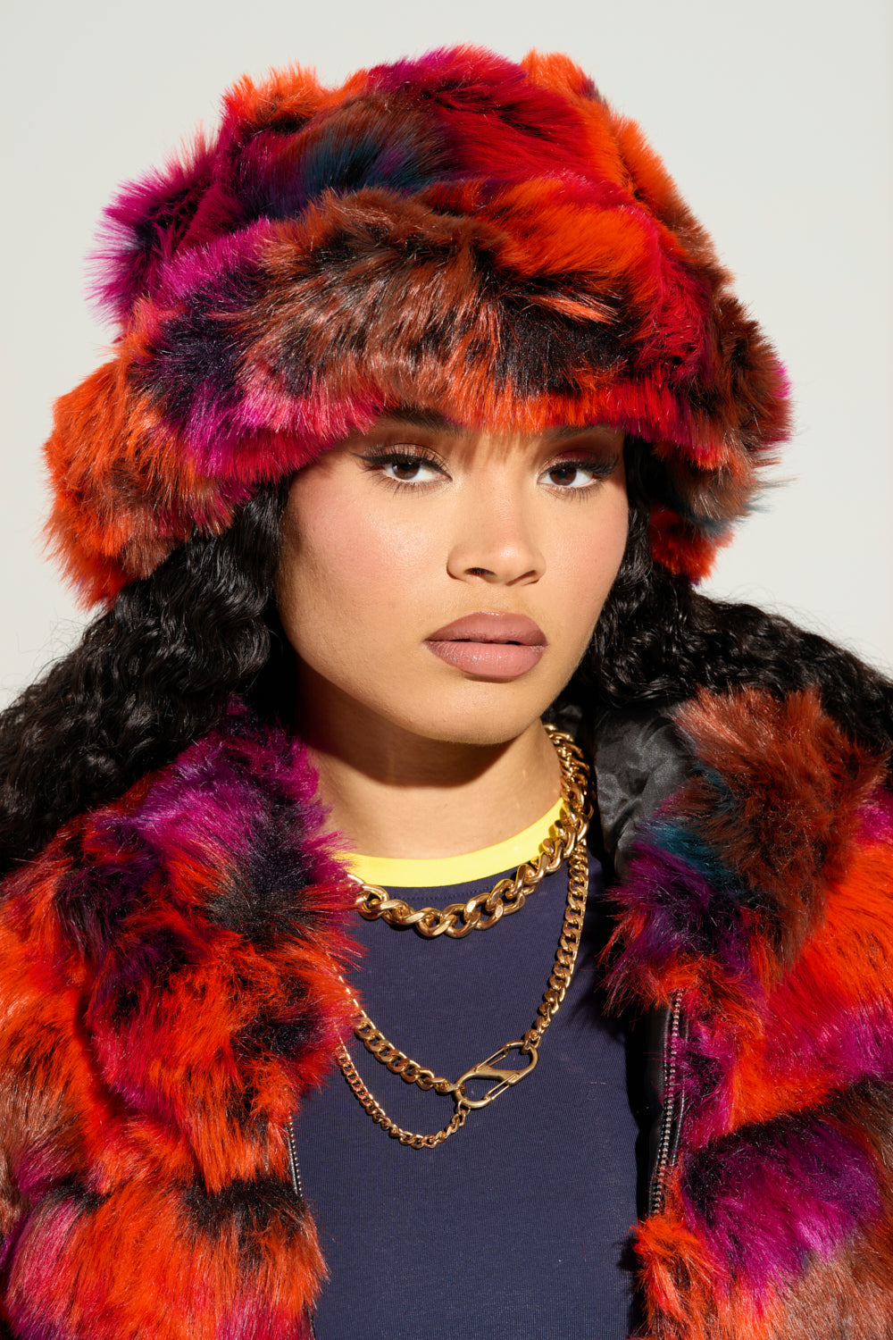 A person in a vibrant multi-colorway fur ensemble flaunts an oversized, floppy silhouette with long curly hair. They maintain a serious expression while wearing layered gold necklaces over a black top. The UPSETTER FAUX FUR BUCKET HAT on their head adds a touch of eclectic charm. 1