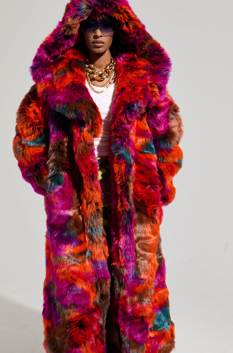Wearing the Upsetter Maxi Faux Fur Coat in Pink Multi, a person displays its vibrant mix of red, purple, and orange hues. With their hood up and sunglasses on, they feature layered necklaces over a crisp white shirt. The plain background ensures their striking fashion choice remains the focal point.