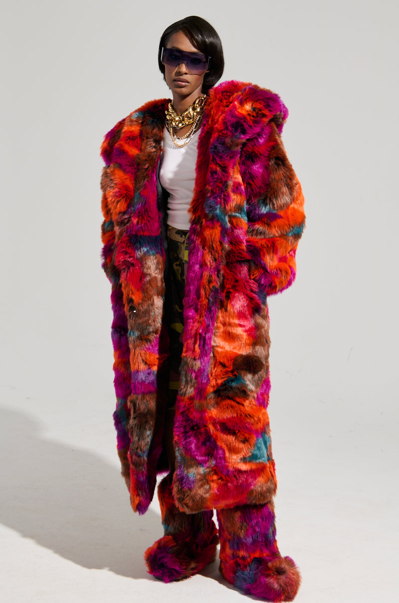 A person confidently poses in an oversized fit, wearing the vibrant UPSETTER MAXI FAUX FUR COAT IN PINK MULTI paired with matching pants. Their ensemble is complemented by a white top, bold sunglasses, a chunky gold necklace, and a short hairstyle, all set against a plain background that highlights the striking colors of their outfit.