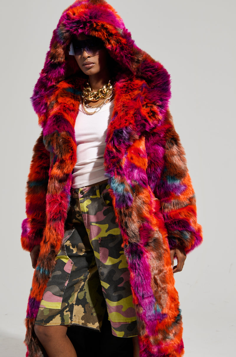 Wearing the UPSETTER MAXI FAUX FUR COAT IN PINK MULTI, this individual complements the vibrant oversized coat with a white top and camouflage skirt. Layered gold necklaces catch the sunlight as they confidently pose, with sunglasses enhancing their chic ensemble.