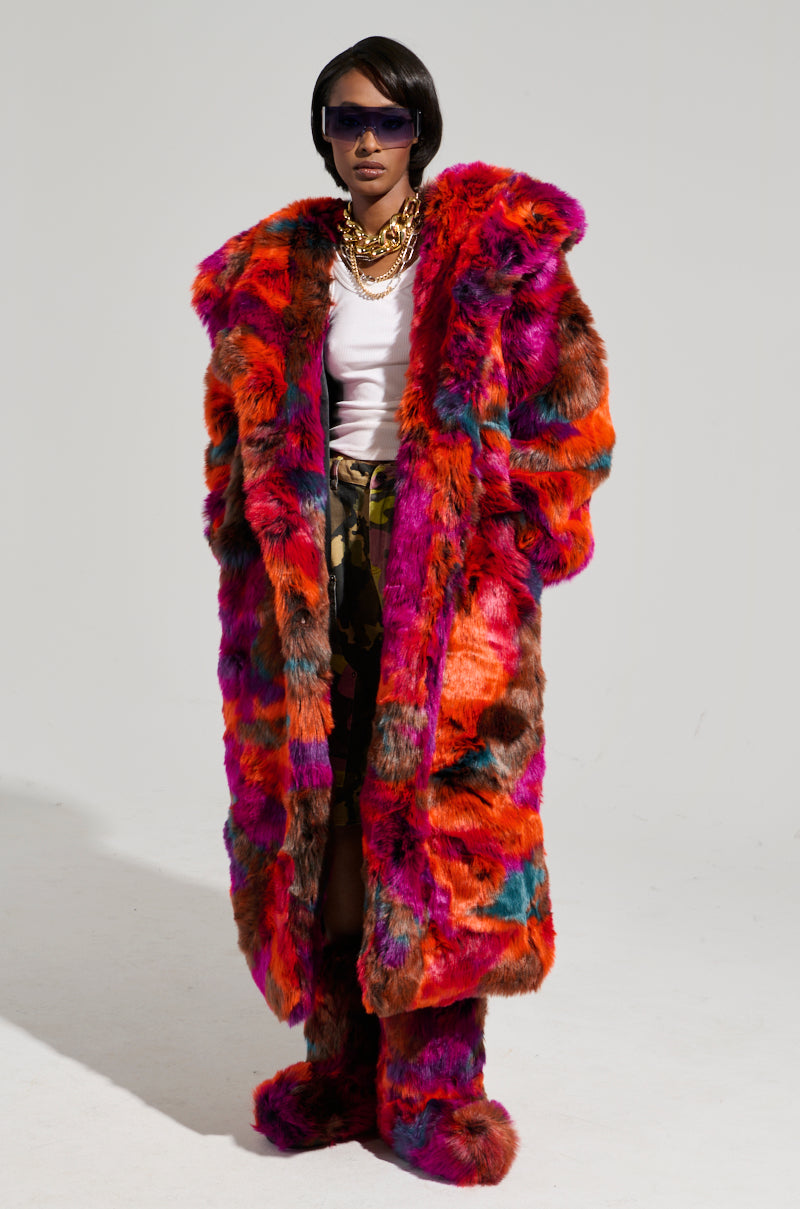 A person stands confidently against a plain background, wearing the UPSETTER MAXI FAUX FUR COAT IN PINK MULTI, matching boots, a white top, and camouflage pants. They accessorize with dark sunglasses and layered gold chains.