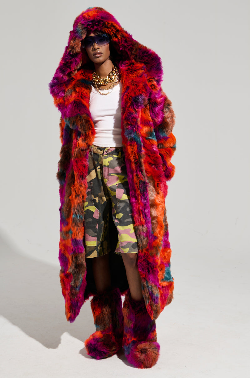 A person sporting the UPSETTER MAXI FAUX FUR COAT IN PINK MULTI, paired with matching boots and camouflage shorts. They’re accessorized with layered necklaces, sunglasses, and a white tank top. The coat's oversized fit and large hood perfectly finish off the bold and eclectic appearance.