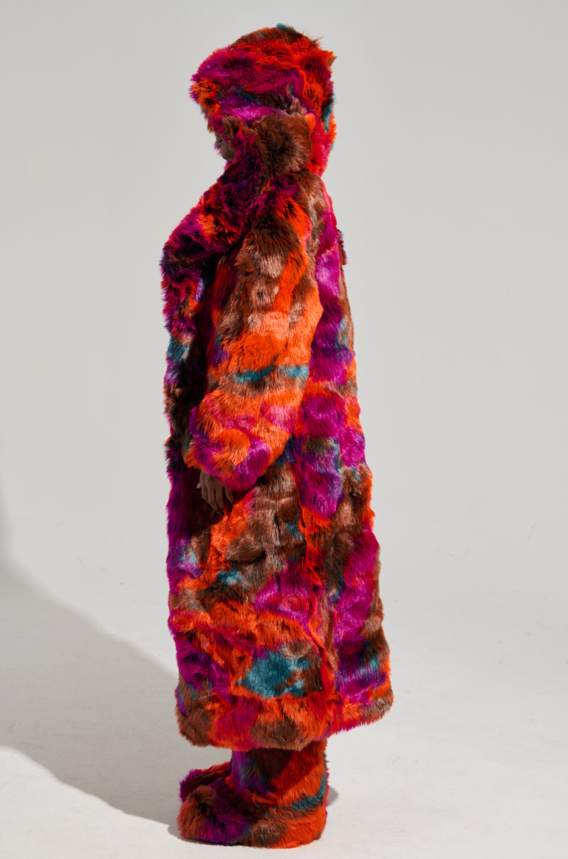 A person wearing the Upsetter Maxi Faux Fur Coat in Pink Multi, featuring an oversized fit and vibrant colorway with a hood, stands in profile against a plain background, complemented by matching boots.