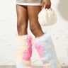 A person wearing the AZALEA WANG UPSETTER PINK STILETTO BOOT, a colorful faux fur knee-high boot in pastel shades of pink, yellow, and blue, holds a small white furry handbag. The outfit features a sheer skirt and a yellow top, set against a light-colored textured wall.