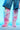 A person showcases the AZALEA WANG UPSETTER PINK STILETTO BOOTS, featuring vibrant knee-highs with a faux fur upper, paired stylishly with denim shorts against a bright blue backdrop. The boots flaunt pointed toes and chic high heels.