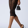 A person strides confidently in black lace leggings and the AZALEA WANG URMI BLACK SANDAL, featuring flower appliques and clear heels, while carrying a small black handbag against a plain white backdrop.