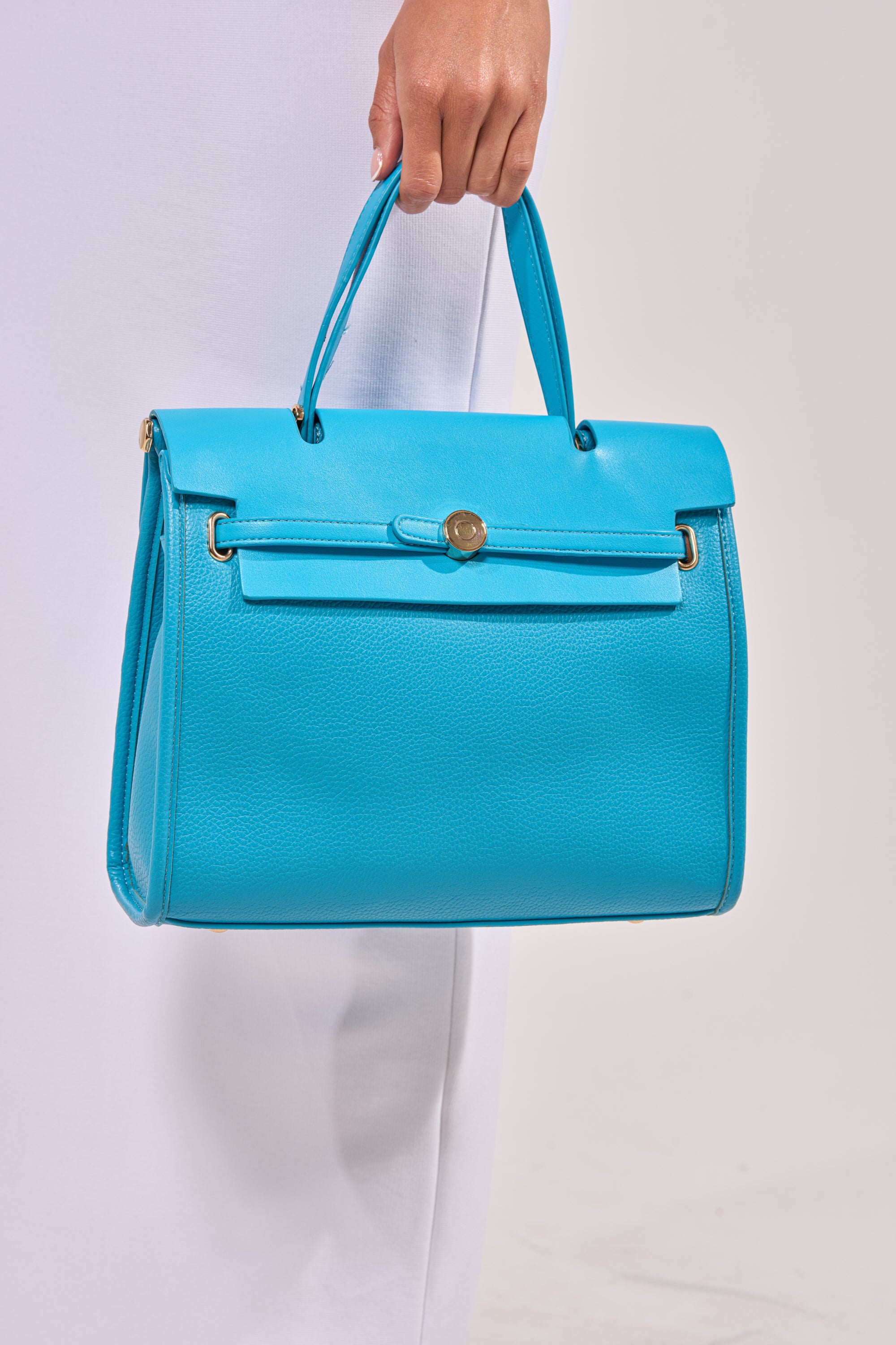 A person holds the TAKE ME OUT TOTE, featuring a bright blue color, top handle, and gold hardware, standing against a plain light background. 1
