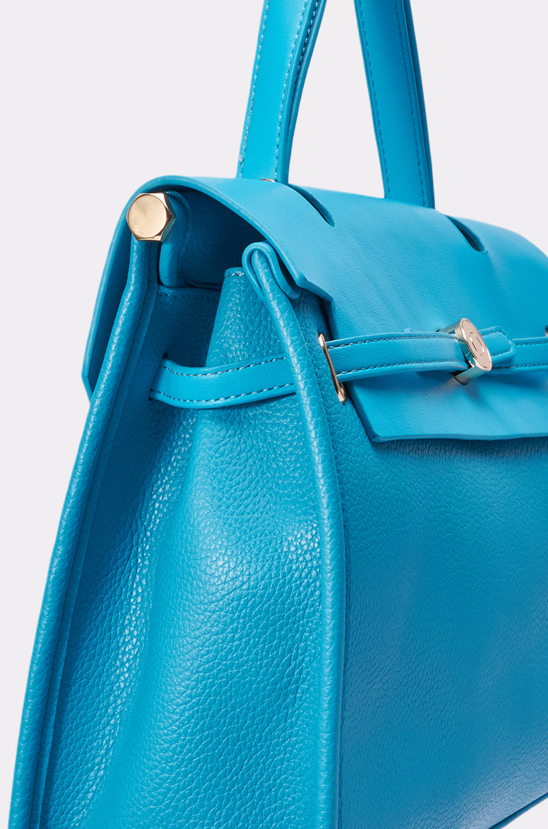 Close-up of the TAKE ME OUT TOTE, a bright blue leather statement bag with gold hardware, textured body, smooth flap, sturdy handle, and crossbody strap set against a plain light background. 3