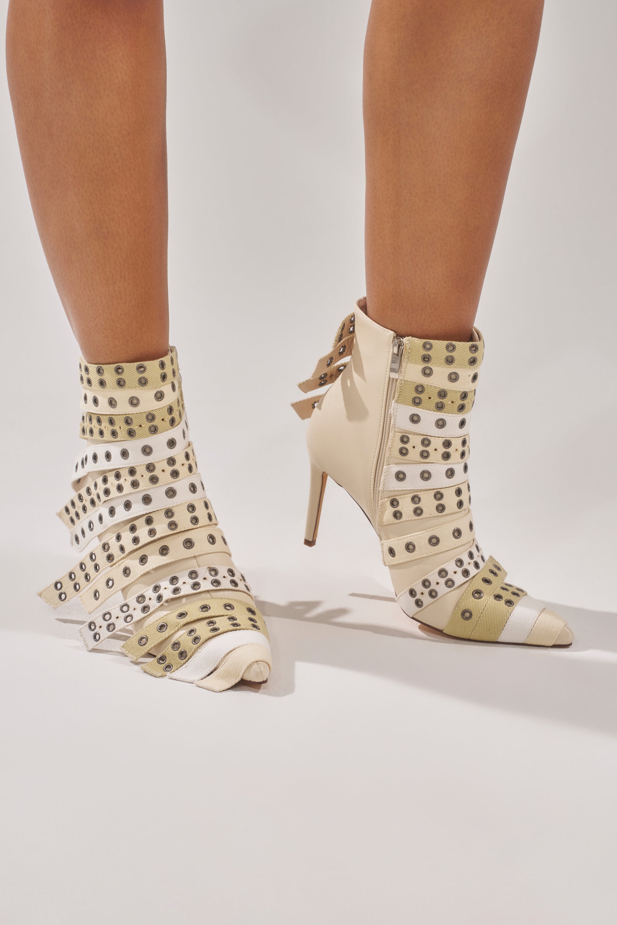 A person wears the AZALEA WANG UZMA Bone Denim Silver Hardware Stiletto Bootie with pointed toes, beige and white striped bands, metallic rivets, silver hardware accents, side zippers, and a slouched layered look on one boot.