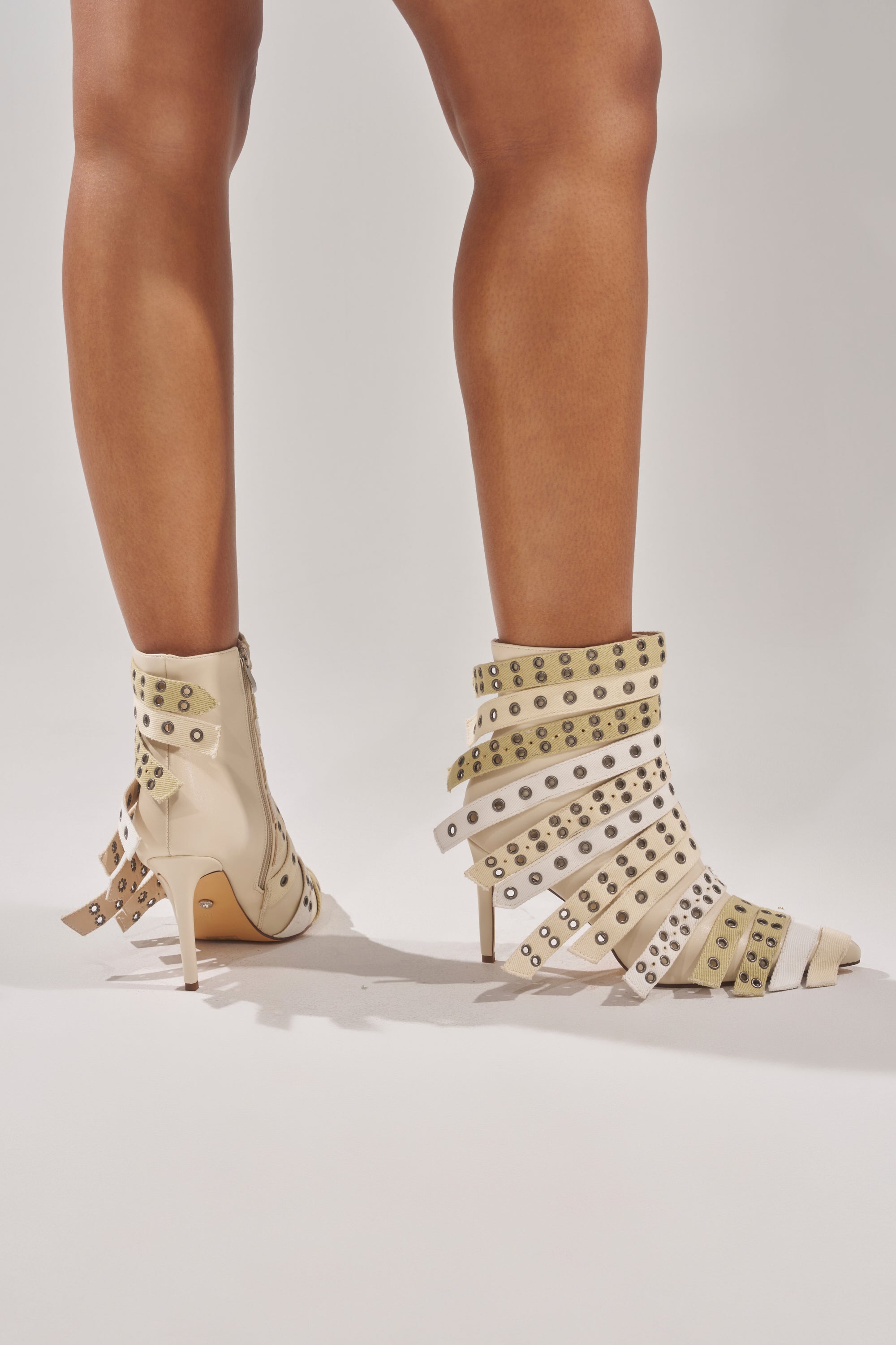 A person wears the AZALEA WANG UZMA BONE DENIM SILVER HARDWARE STILETTO BOOTIE, featuring high heels, pointed toes, and beige and white straps with silver hardware, standing against a plain light background.